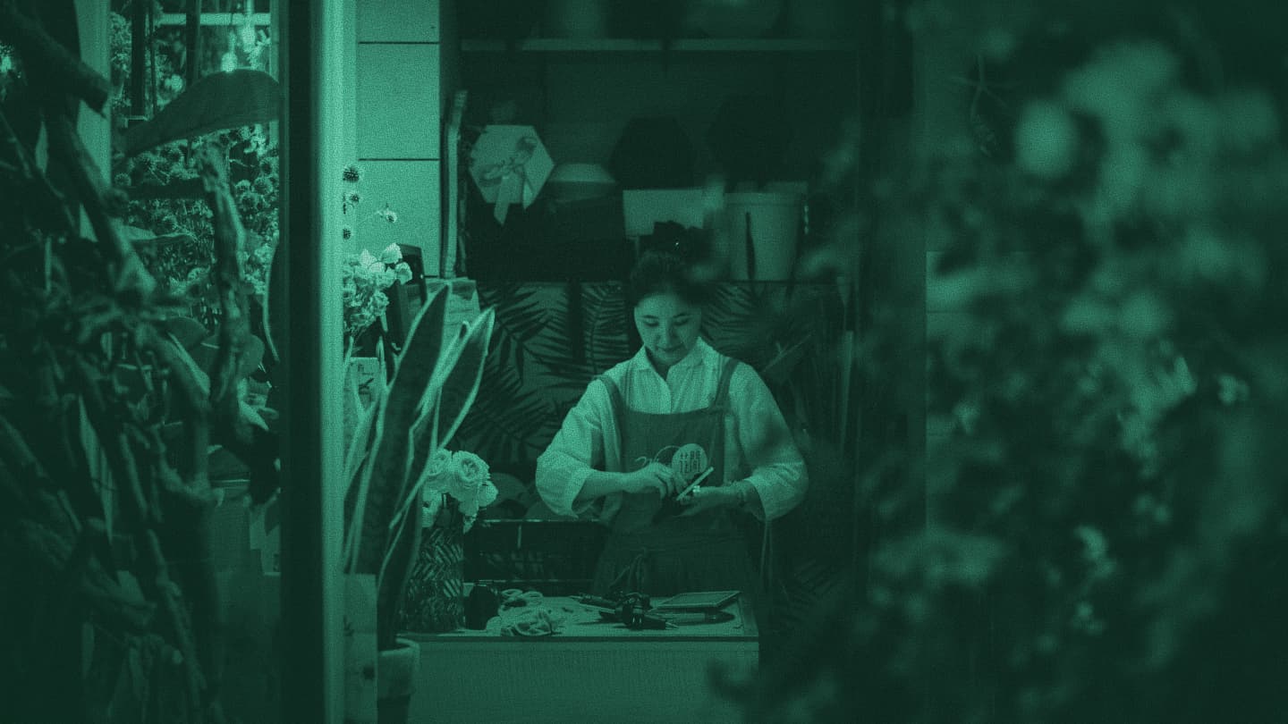 Woman in an apron working at a counter in a plant shop, viewed through foliage, teal tint