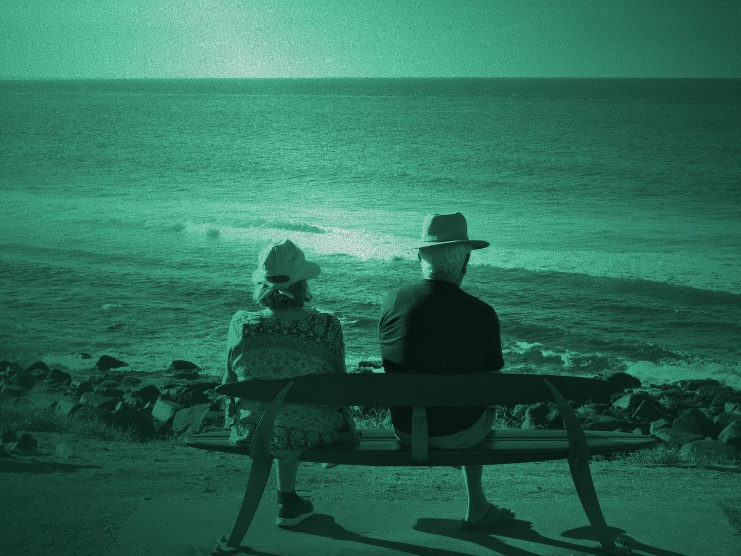 Older couple sitting on a park bench looking out to sea, teal monochrome photo