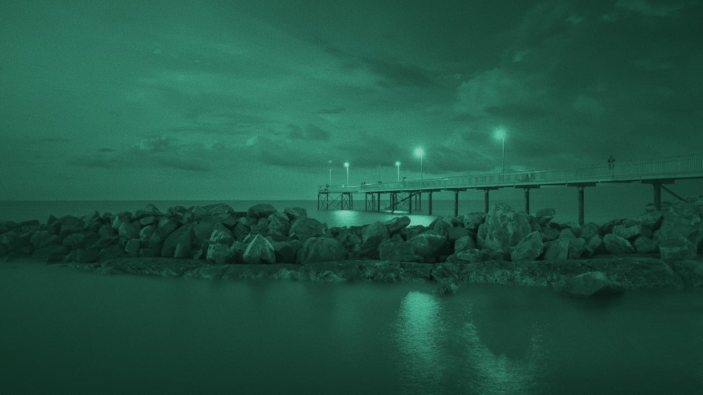 Long-exposure night photo of a lit pier over calm water behind a rocky breakwater, teal tint